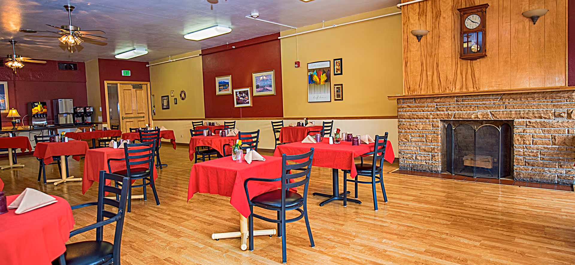 A dining room with multiple tables covered in red tablecloths and black chairs. Each table is set with napkins and small flower vases. The room has wooden flooring, a stone fireplace with a wooden mantle and a clock above it, and walls painted in red and yellow. Ceiling fans and fluorescent lights are visible on the ceiling, and there is a beverage station with coffee dispensers and a refrigerator in the background.