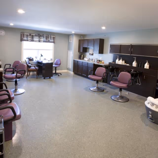 Interior view of a salon area in a senior living facility with multiple purple salon chairs, hair washing stations with shampoo bottles, cabinets along the wall, and a desk with chairs near a window with a patterned valance.