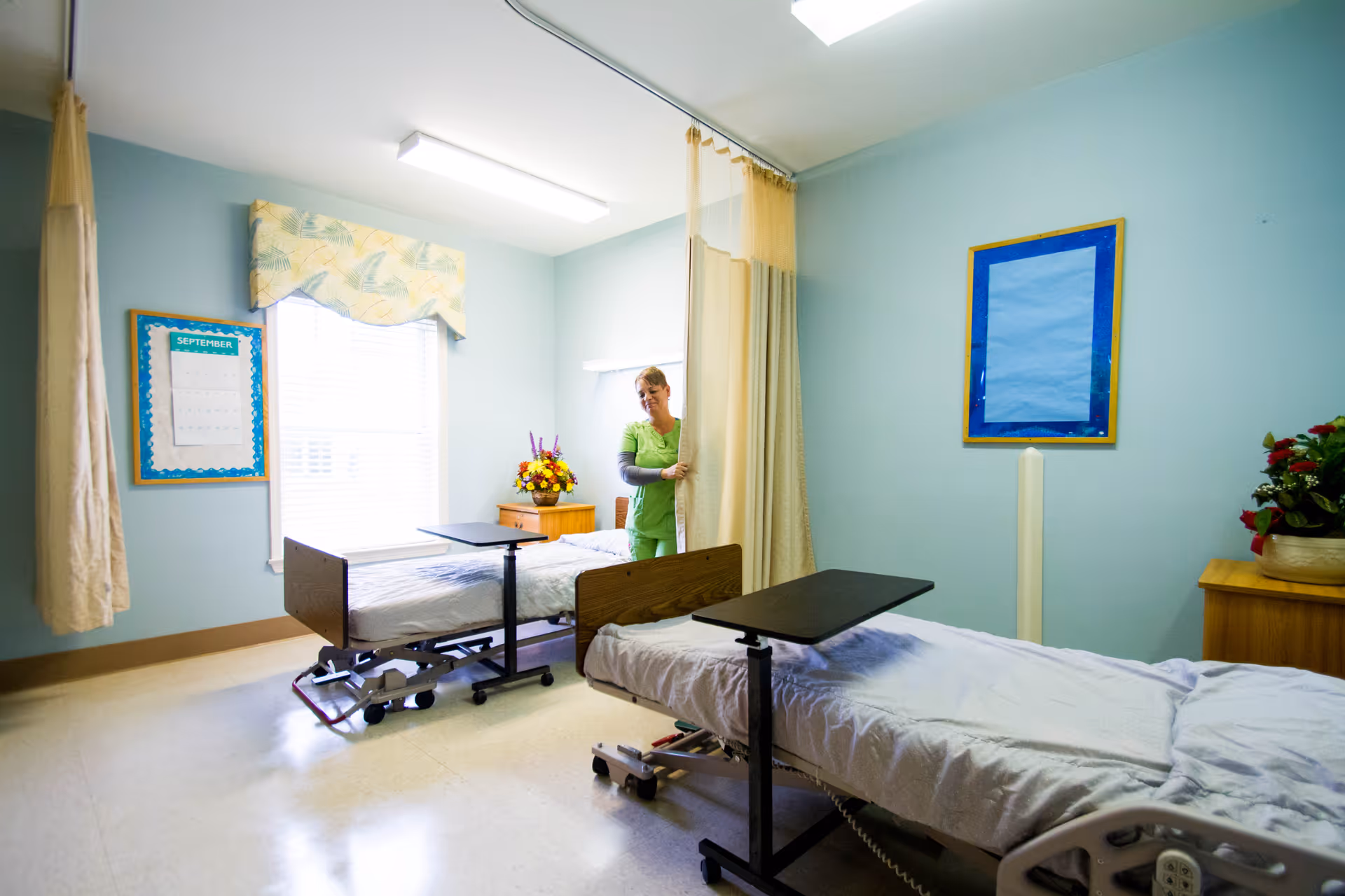 A hospital or nursing center room with two beds separated by a privacy curtain. A healthcare worker in green scrubs is adjusting the curtain. The room has light blue walls, a window with a valance, a calendar on the wall, and flower arrangements on wooden bedside tables.