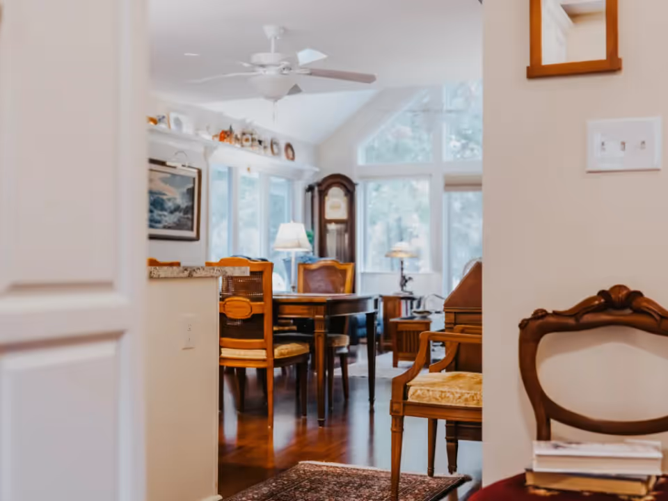 View through a doorway into a bright interior showing a dining table with wooden chairs, a ceiling fan, lamps, and large windows.