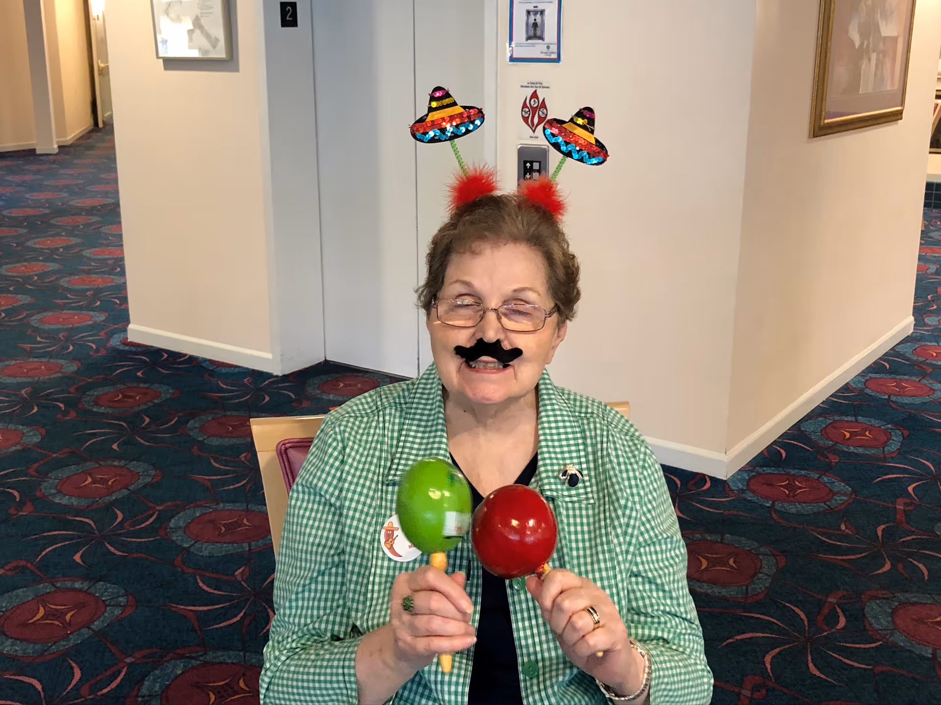 An elderly woman wearing glasses and a green checkered shirt is sitting indoors on a chair. She has playful digital decorations added to her image, including a black mustache, red fluffy antennae with colorful sombrero hats, and is holding a green and a red maraca. The background shows a hallway with blue and red patterned carpet and white walls.
