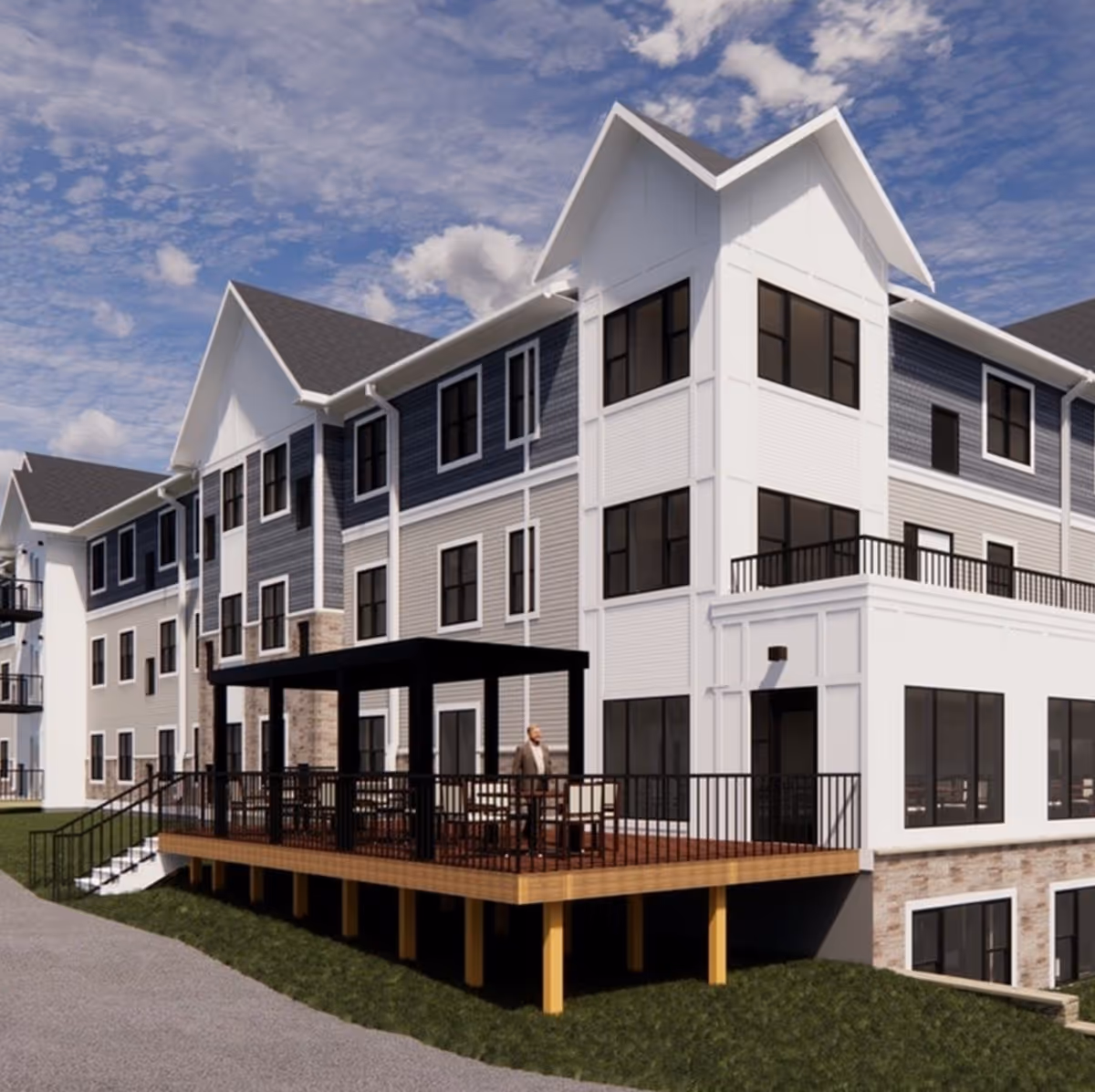 Exterior view of a multi-story senior living facility building with a large wooden deck featuring a pergola and outdoor seating. The building has a combination of white and gray siding with multiple windows and a sloped roof under a partly cloudy sky.