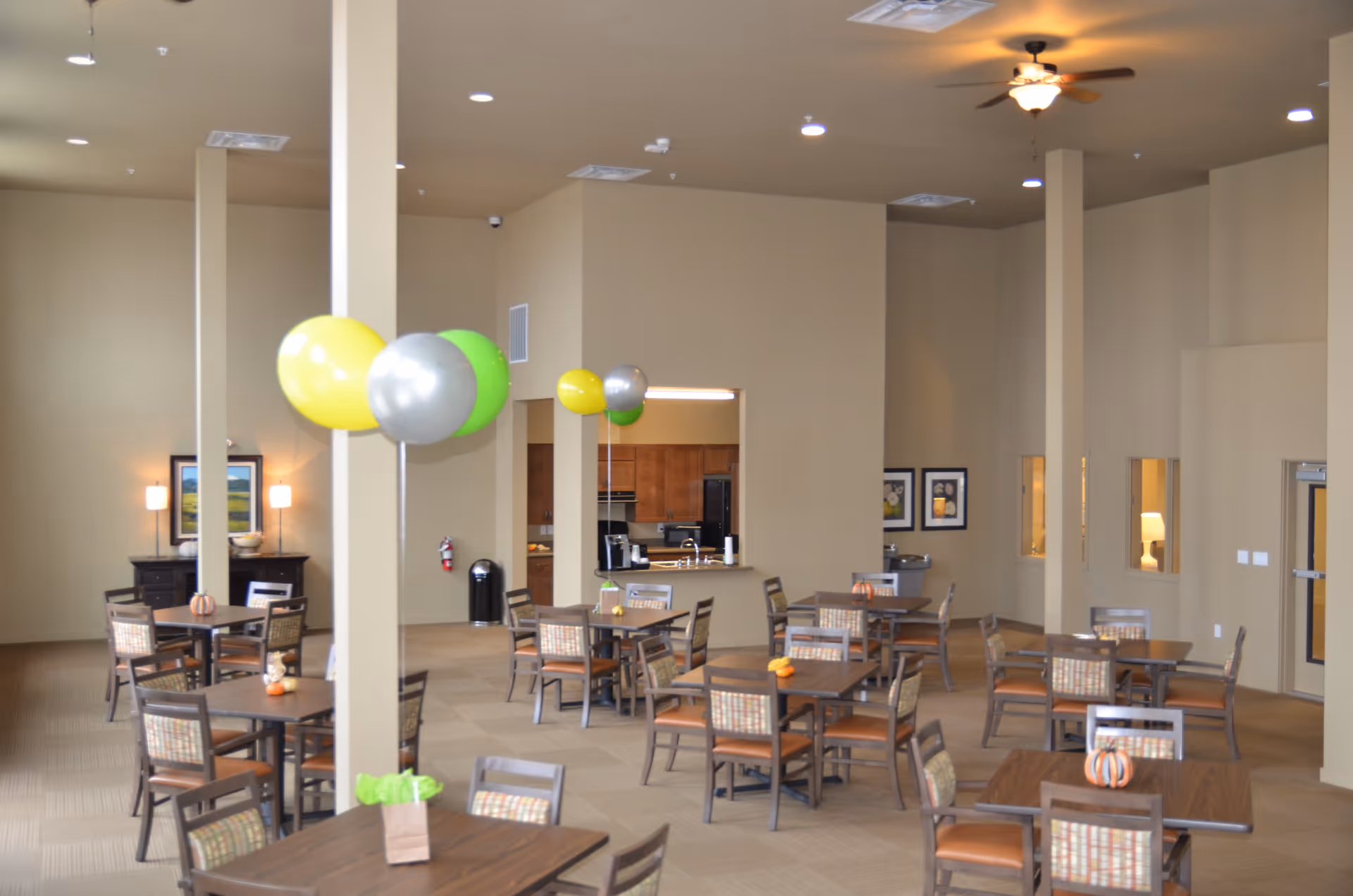 A spacious dining room in a senior living facility with multiple wooden tables and chairs arranged neatly. The room has beige walls and carpeted floors, decorated with small pumpkins on the tables and colorful balloons tied to a pillar. There is a ceiling fan with a light fixture, framed artwork on the walls, and a kitchen area visible through a pass-through window.