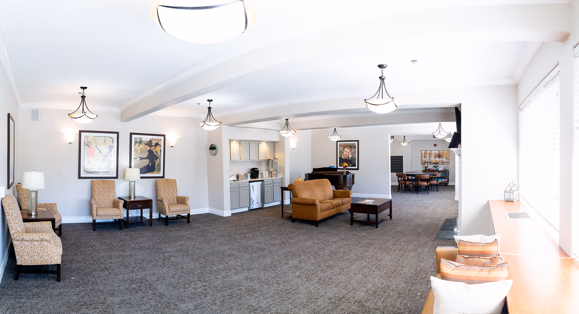 A spacious senior living common area with beige walls and carpeted floor. The room features multiple seating options including patterned armchairs, a brown loveseat, and a window bench with cushions. There are several ceiling light fixtures and framed artwork on the walls. A small kitchenette area with cabinets, a coffee maker, and a mini fridge is visible. In the background, there is a piano and a dining area with a round table and chairs.