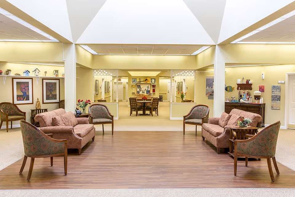 Bright common lounge in a senior living facility with upholstered sofas and chairs arranged around a central wood floor and a dining area beyond.