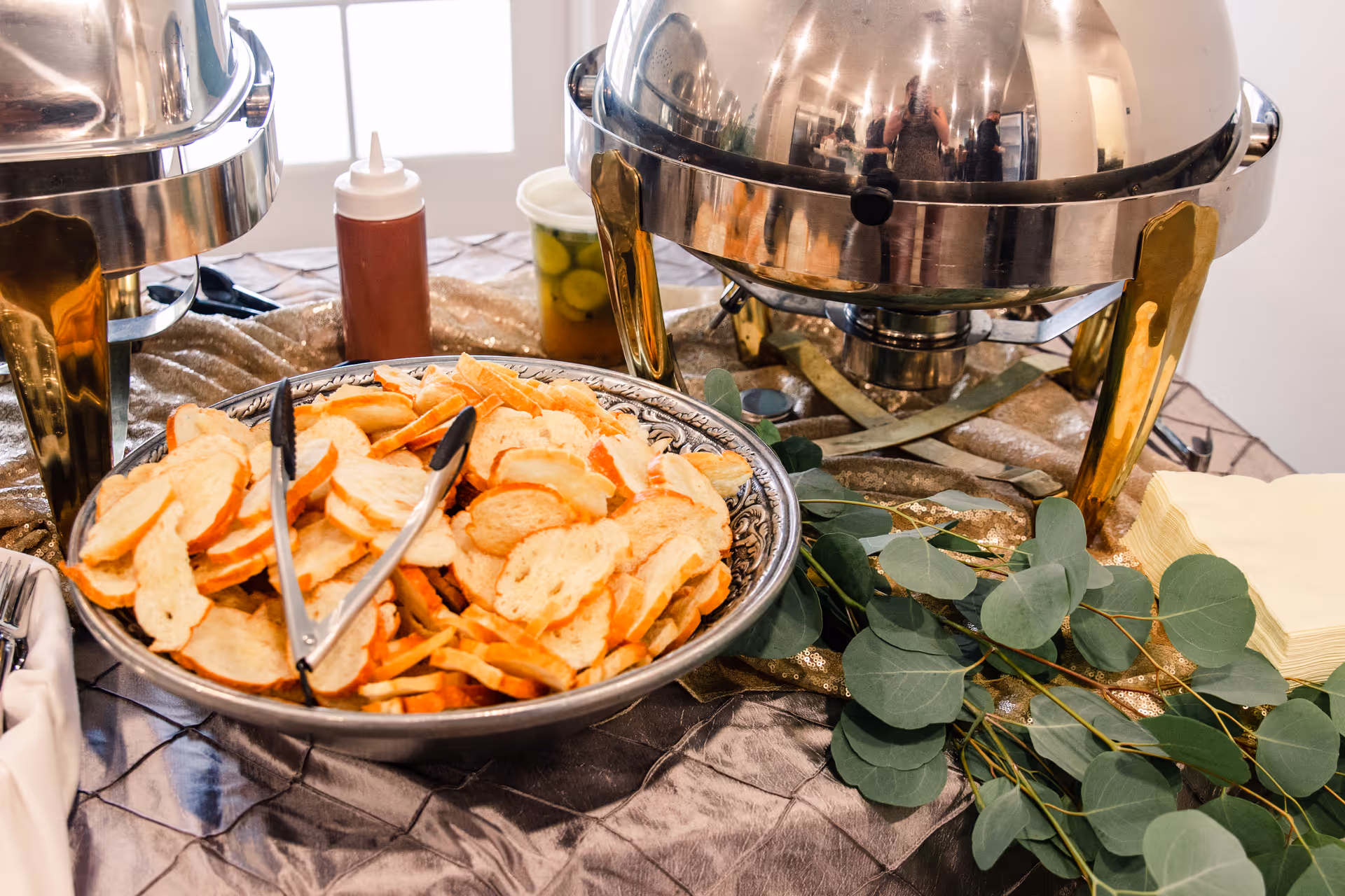 Buffet table with a bowl of sliced bread, chafing dishes, tongs, condiments and eucalyptus sprigs.