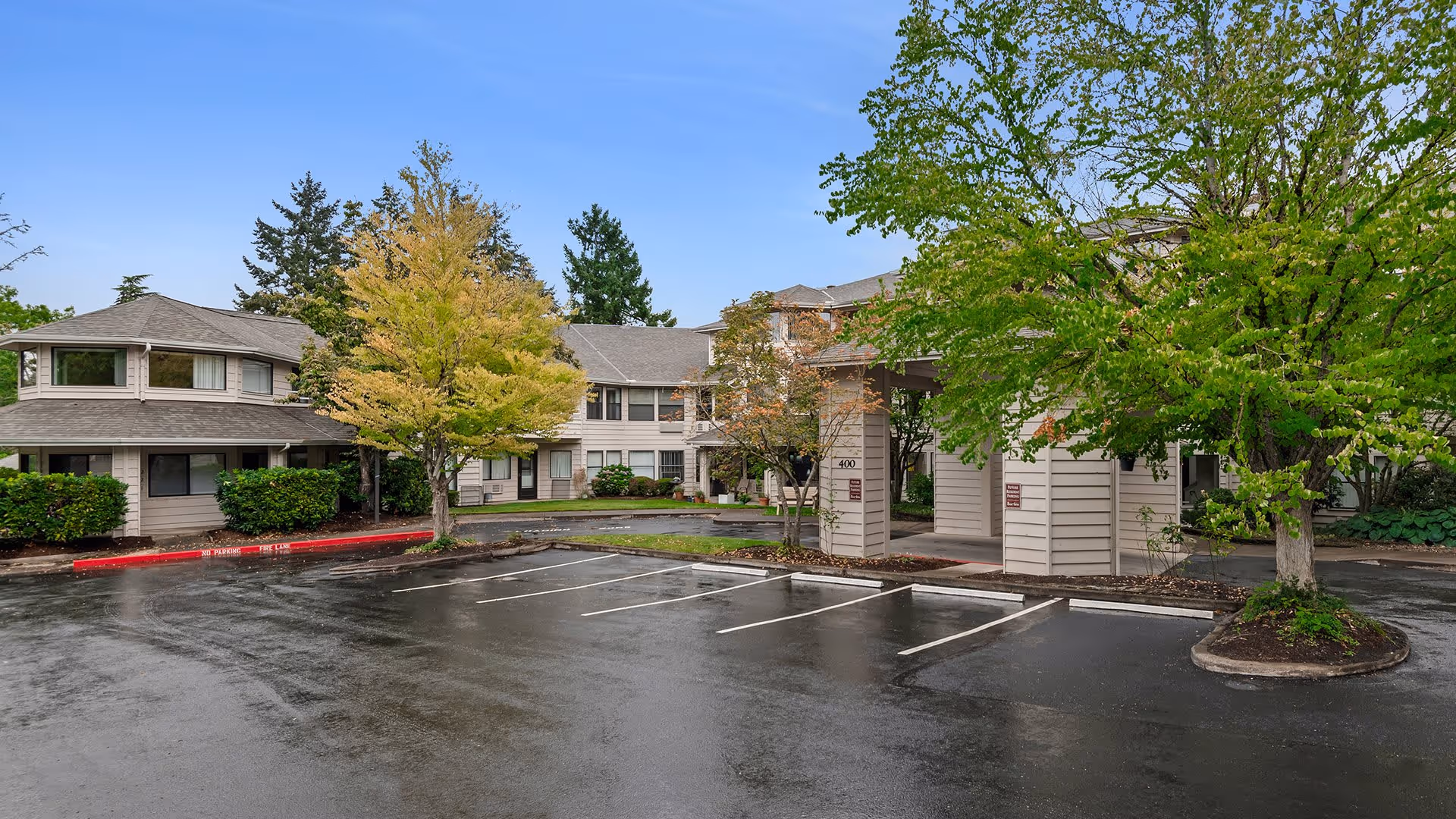 Exterior view of Holiday Hidden Lakes facility showing a two-story building with beige siding surrounded by trees with green and yellow leaves. There is a covered entrance with pillars and an empty parking lot in front on a wet pavement under a clear blue sky.