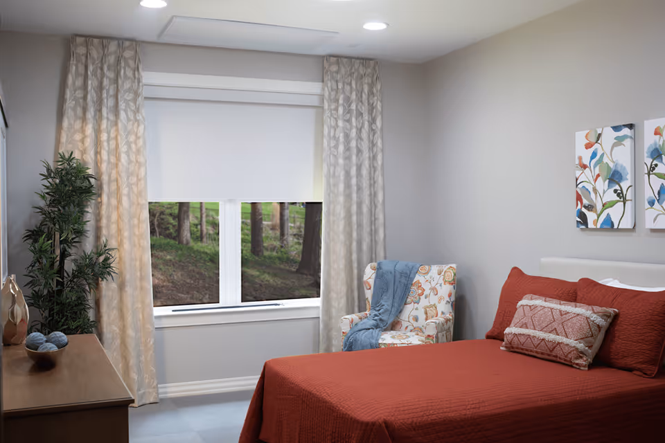 A cozy bedroom with a bed covered in a red quilt and matching pillows, a floral patterned armchair with a blue throw blanket, a wooden dresser with decorative items, beige curtains framing a window showing a view of trees outside, and two colorful floral paintings on the wall.