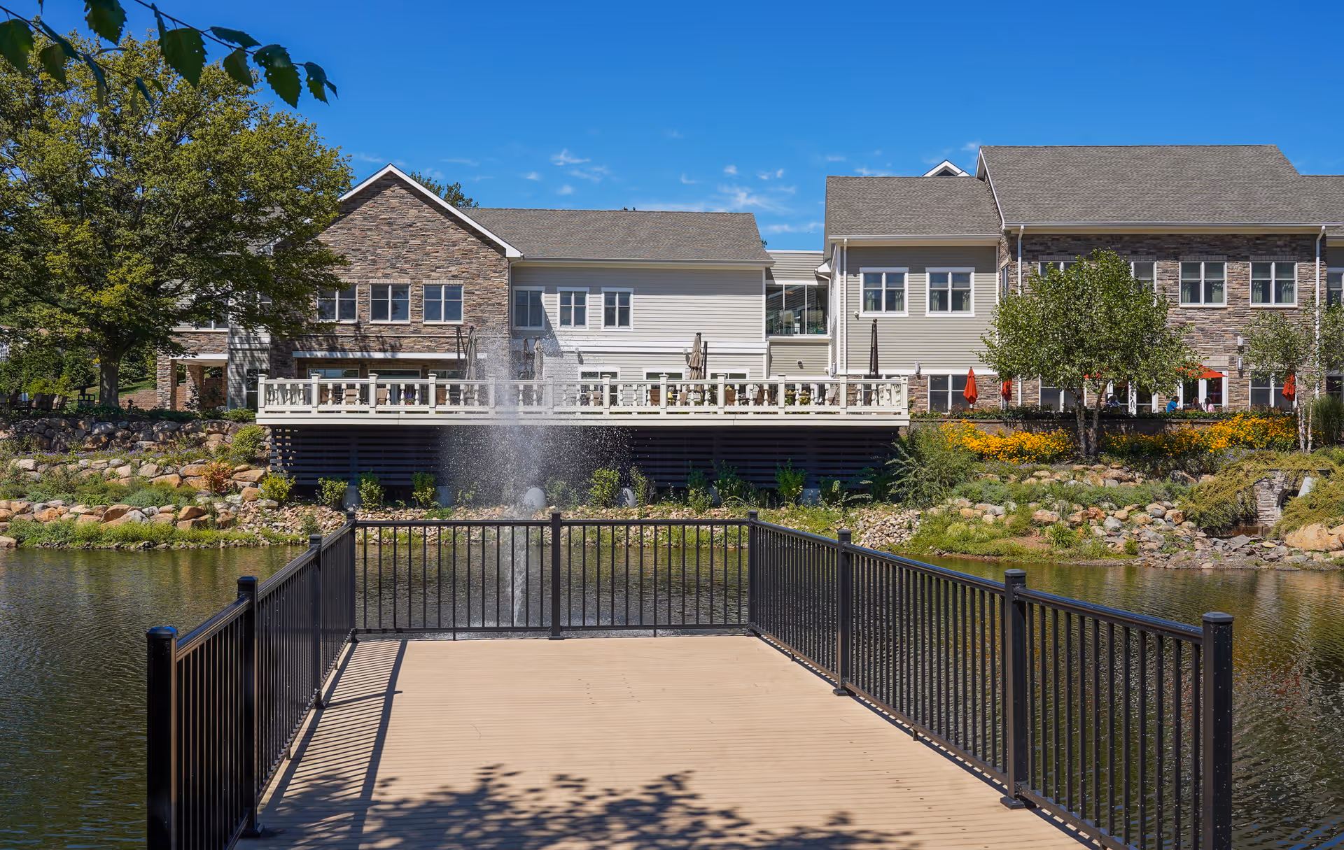 View of a senior living facility named Pine Run Village with a large pond in the foreground, a wooden dock with black railings extending into the pond, and a water fountain spraying water in the center of the pond. The building has a combination of stone and light-colored siding with multiple windows, surrounded by trees and landscaped greenery under a clear blue sky.