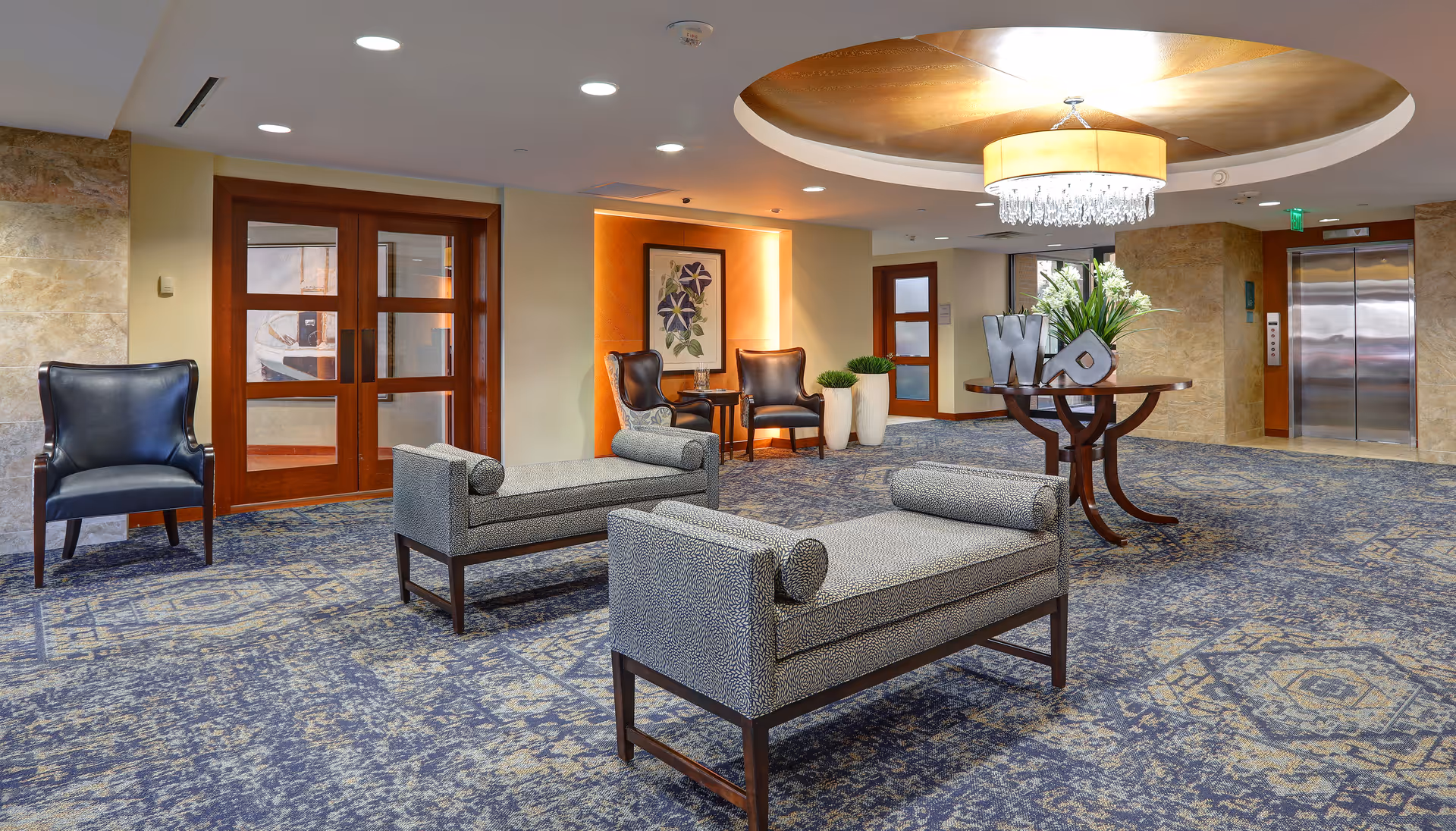 A spacious, well-lit lobby with upholstered benches and chairs, a decorative round table with plants, and an elevator in the background.