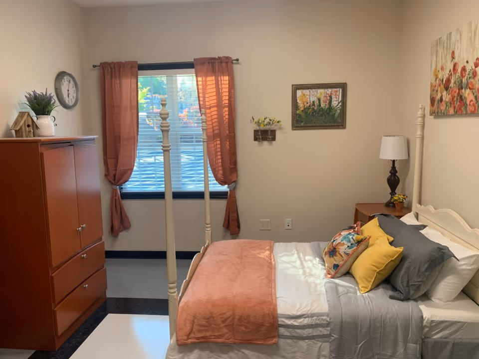 A cozy bedroom with a white four-poster bed made with white, gray, and peach bedding and colorful pillows. There is a wooden nightstand with a lamp and small flower arrangement beside the bed. A wooden dresser with a plant and a small birdhouse on top is positioned near a window with peach-colored curtains. The walls are decorated with two floral paintings.