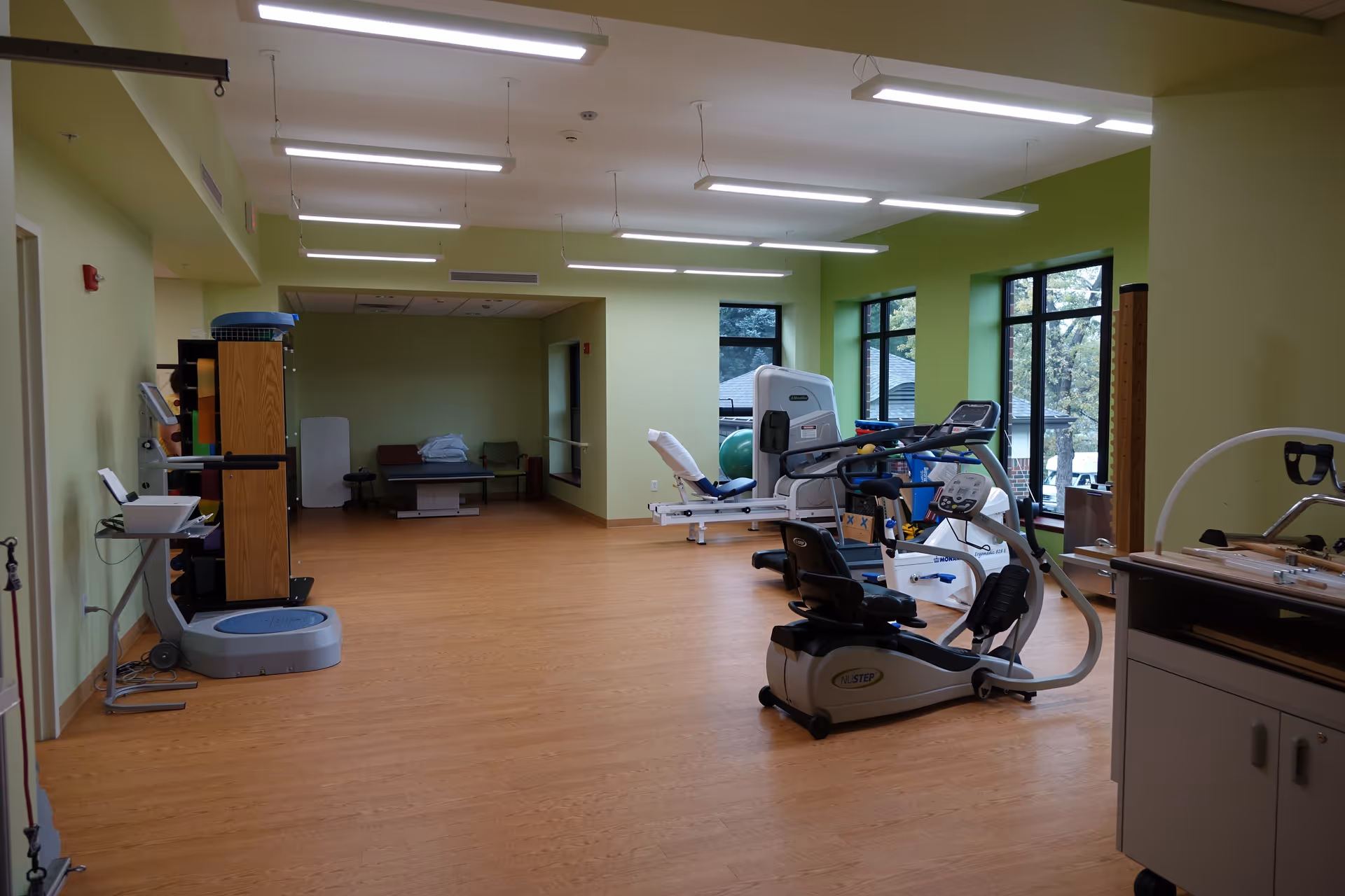 A spacious room with light green walls and wooden flooring containing various exercise and rehabilitation equipment including a recumbent exercise bike, a leg press machine, and therapy tables. Large windows on the right side let in natural light and show trees outside.