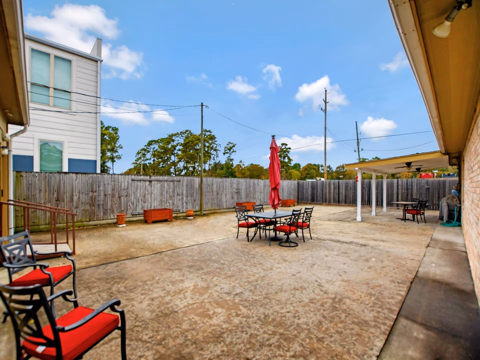 Outdoor patio area with concrete flooring, several black metal chairs with red cushions, a table with a closed red umbrella, a covered seating area with ceiling fans, and a wooden privacy fence surrounding the space under a blue sky with some clouds.