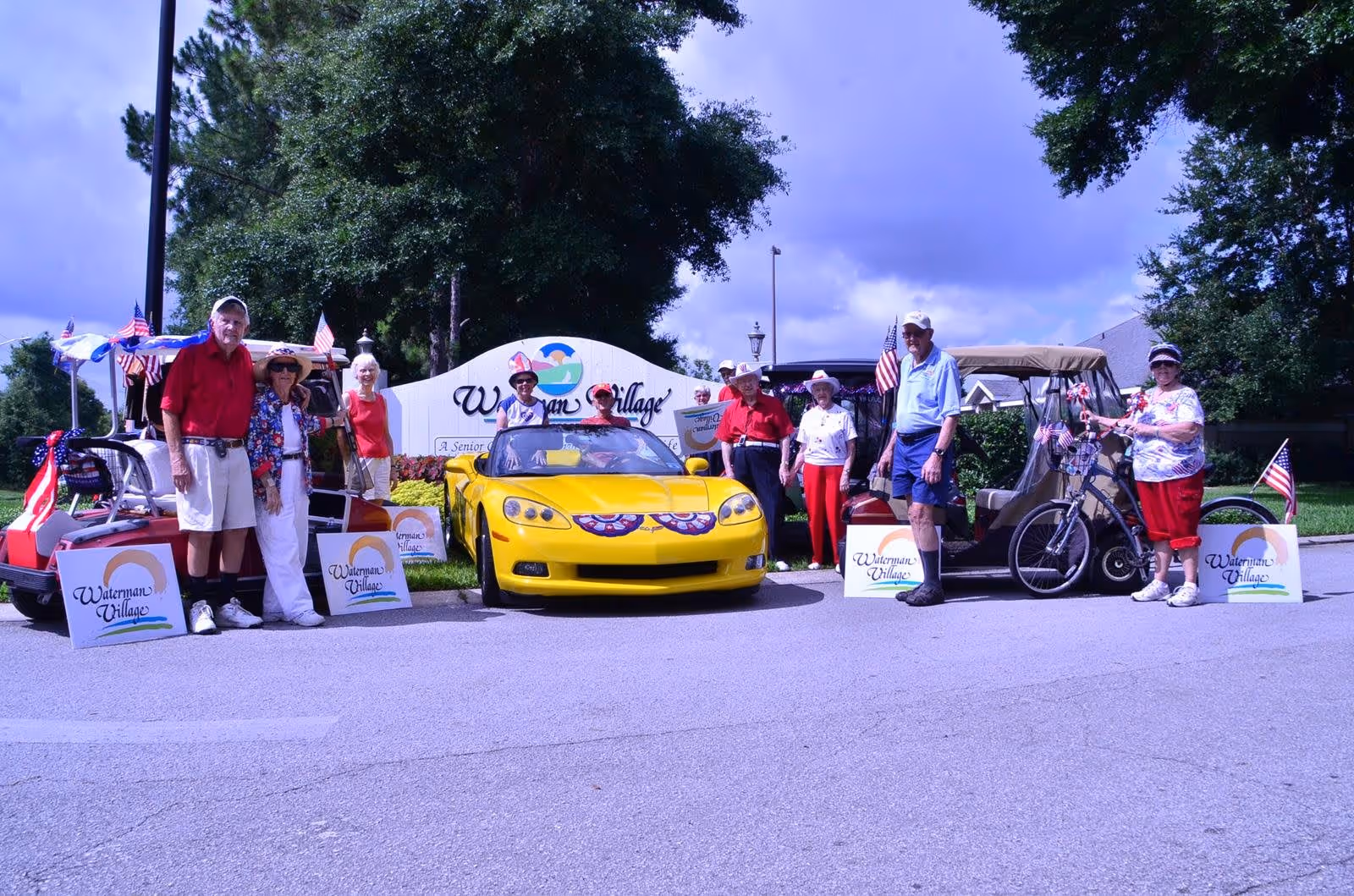 A group of elderly people standing outdoors on a paved road in front of a Waterman Village sign. They are posing with a yellow convertible car decorated with patriotic bunting and two golf carts adorned with American flags. The scene is set on a sunny day with trees and a partly cloudy sky in the background.