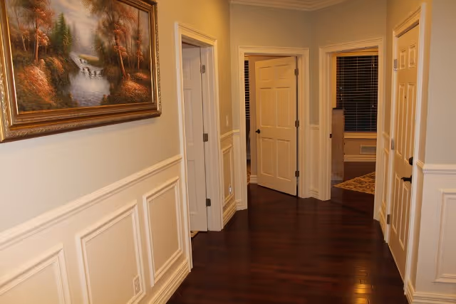 A hallway with dark wooden floors and cream-colored walls featuring decorative wainscoting. There are multiple white doors along the hallway, one of which is open, revealing a room with a window and blinds. A framed landscape painting hangs on the left wall.