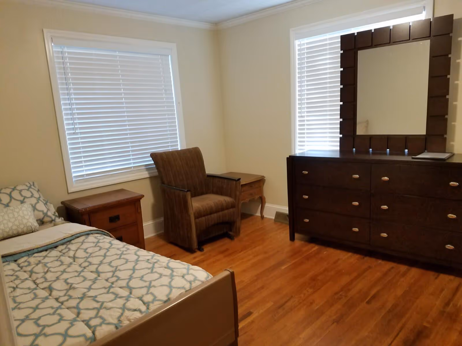 A bedroom with a single bed covered in a patterned comforter, a wooden nightstand, a brown upholstered armchair, a wooden dresser with a large mirror, and two windows with white blinds.