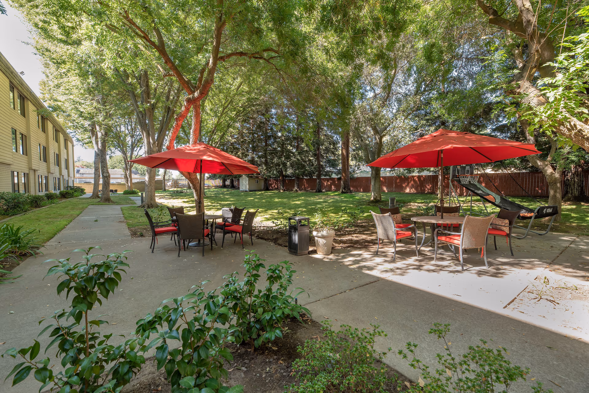 Outdoor patio area at Campus Commons Senior Living with two round tables, each shaded by a red umbrella, surrounded by chairs with red cushions. The patio is set on a concrete surface with green grass, trees, and shrubs around. A building is visible on the left side, and a basketball shooting game is on the right side under the trees.