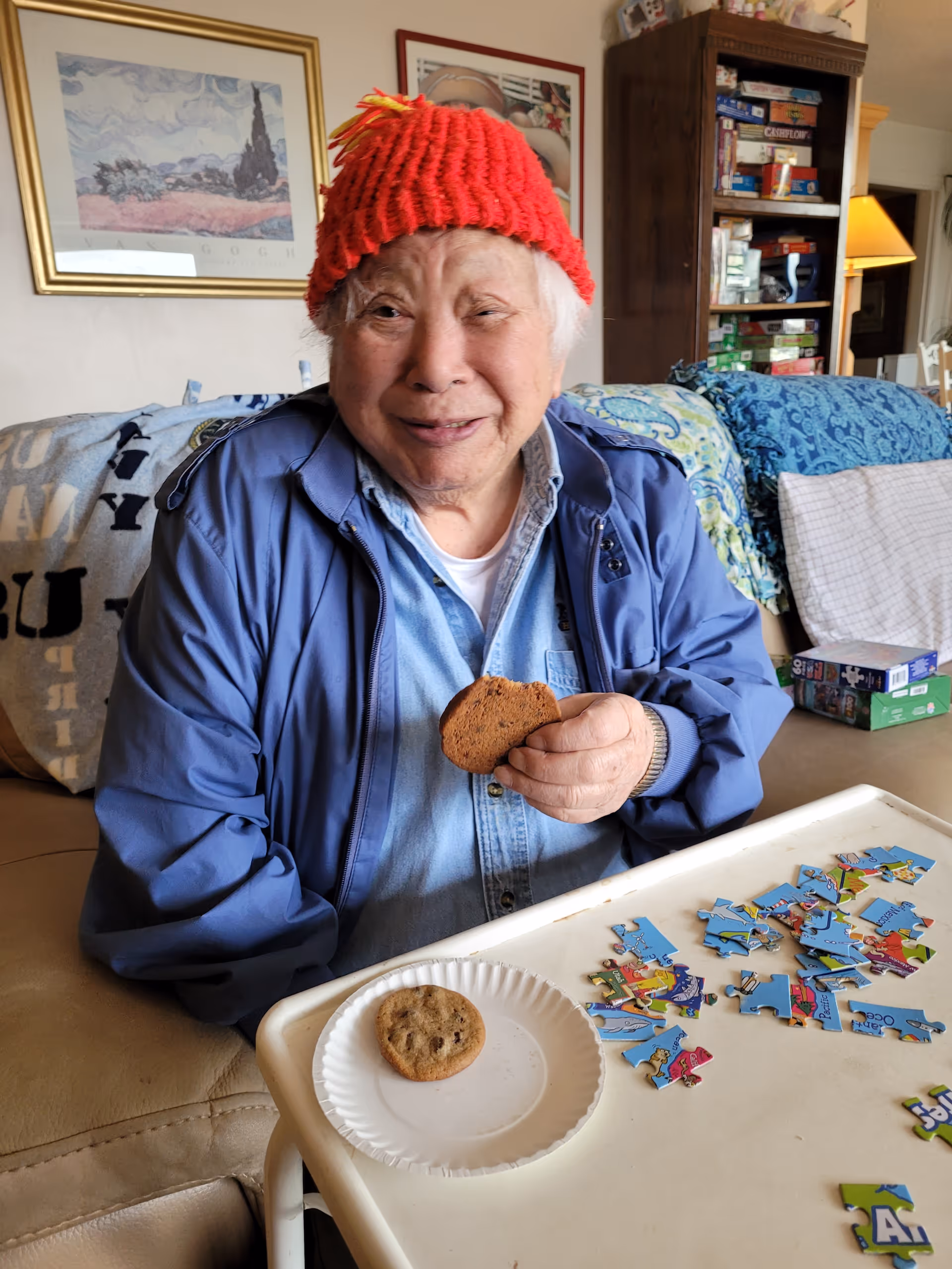 An elderly person wearing a red knit hat and blue jacket is sitting on a couch, holding a chocolate chip cookie. In front of them is a tray with a paper plate holding another cookie and several scattered puzzle pieces. Behind them, there is a bookshelf filled with board games and framed artwork on the wall.