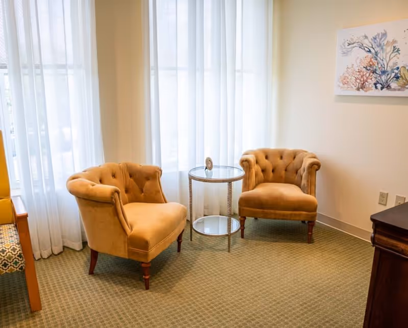 Two tufted beige armchairs flanking a small glass side table in a softly lit sitting area with sheer curtains and wall art.