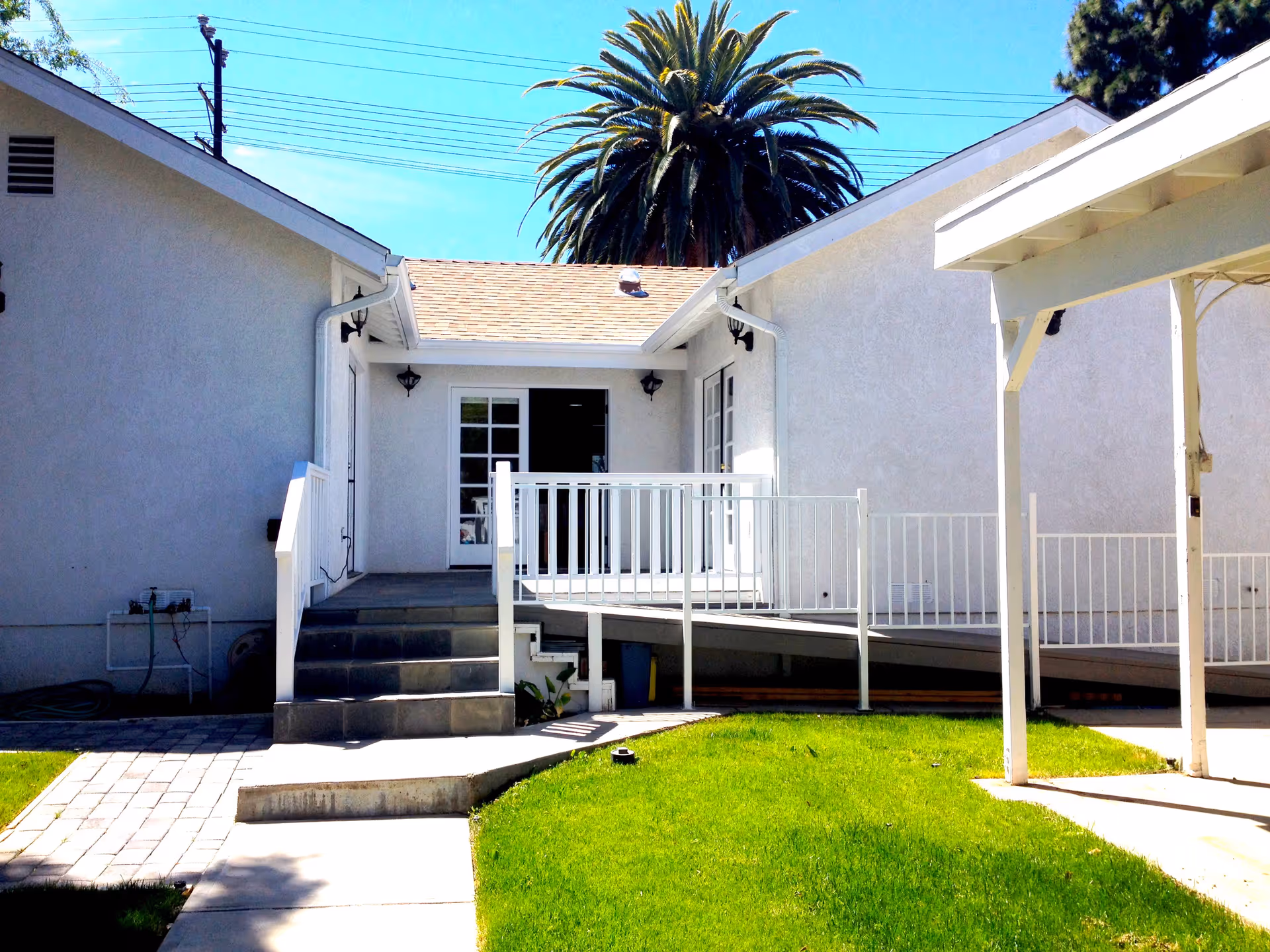Exterior view of a white building with a small staircase and a wheelchair ramp leading to a door. There is a green lawn in the foreground and a large palm tree visible behind the building under a clear blue sky.