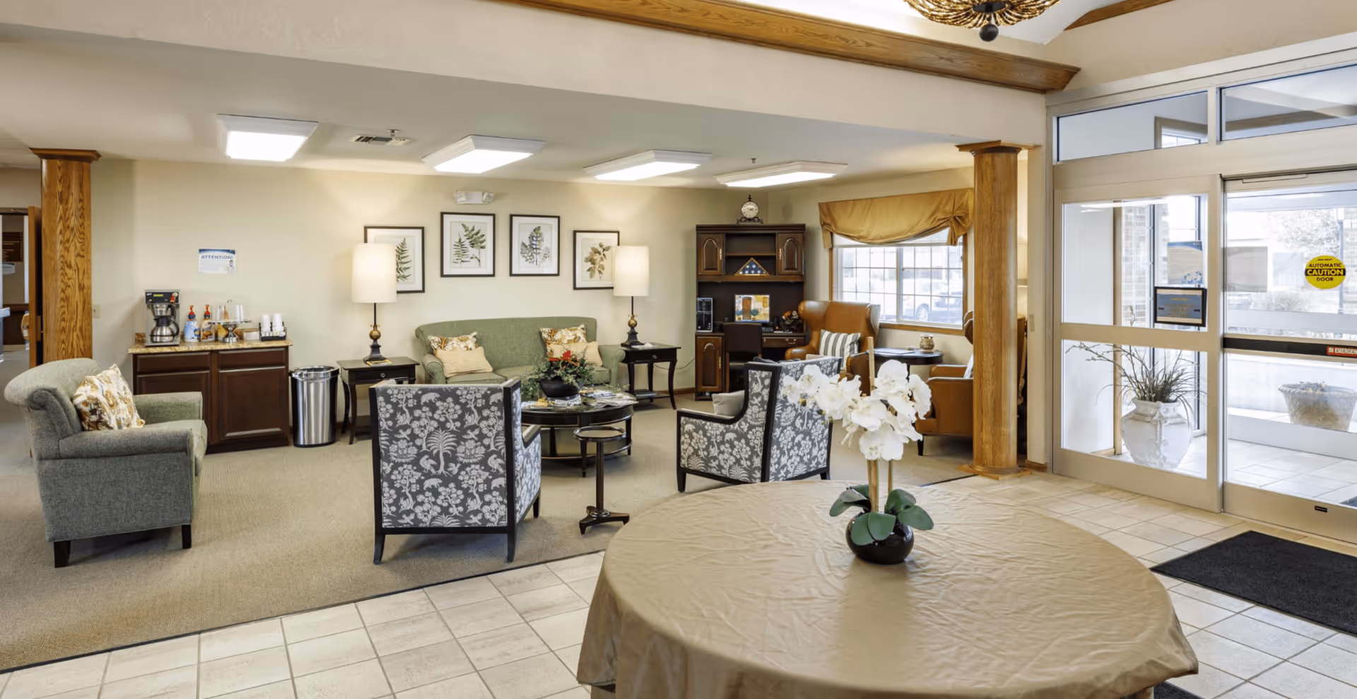 Reception lounge with upholstered chairs and sofas, a round table topped with flowers, and glass entry doors.