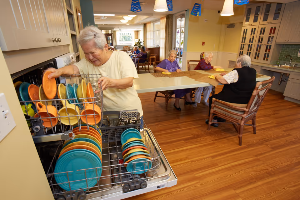 An elderly woman is unloading colorful plates from a dishwasher in a kitchen area. In the background, three elderly women are sitting around a table, cleaning or wiping the table surface. The room has wooden flooring, light yellow walls, and cabinets with glass doors.