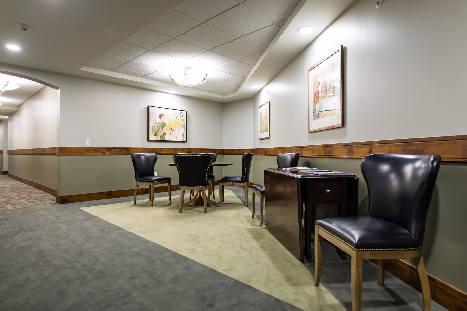 A seating area in a hallway with four black leather chairs arranged around a small round table on a light green carpet. There is a dark wooden side table with magazines on it and three framed paintings on the walls. The walls are painted light gray with a wooden chair rail and green lower paneling. The ceiling has recessed lighting and a central light fixture.