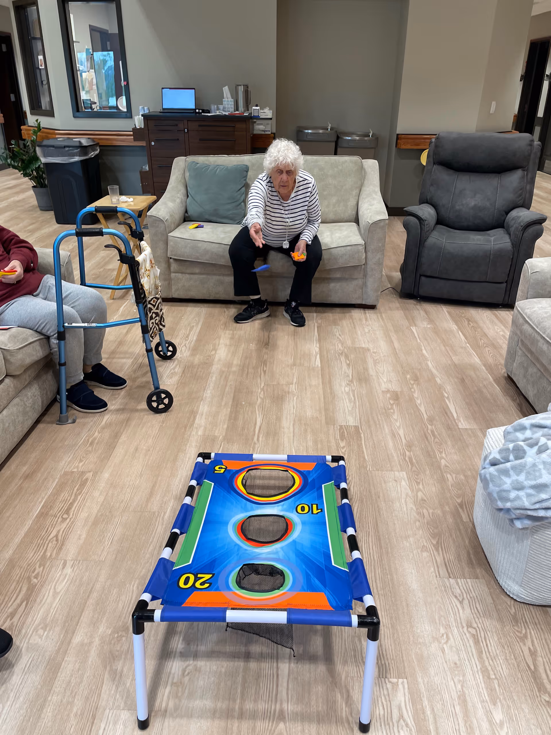 An elderly woman sitting on a beige couch in a living room area, playing a bean bag toss game. There is a walker nearby and another person partially visible sitting on a couch to the left. The room has light wood flooring, a gray recliner chair, and a cabinet with a water dispenser in the background.
