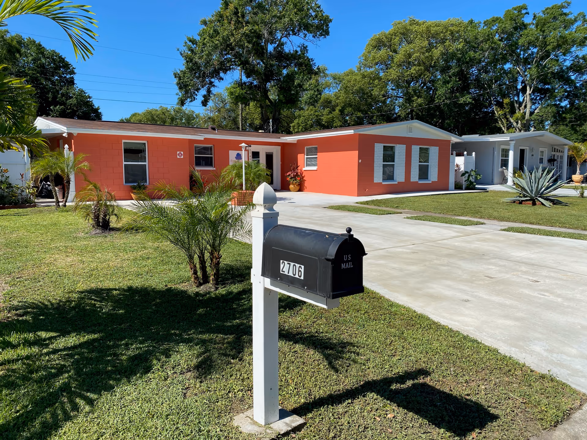 A single-story coral-colored building with white trim surrounded by a well-maintained lawn and palm plants. In the foreground, there is a black mailbox on a white post with the number 2706. The sky is clear and blue, and there are large trees in the background.