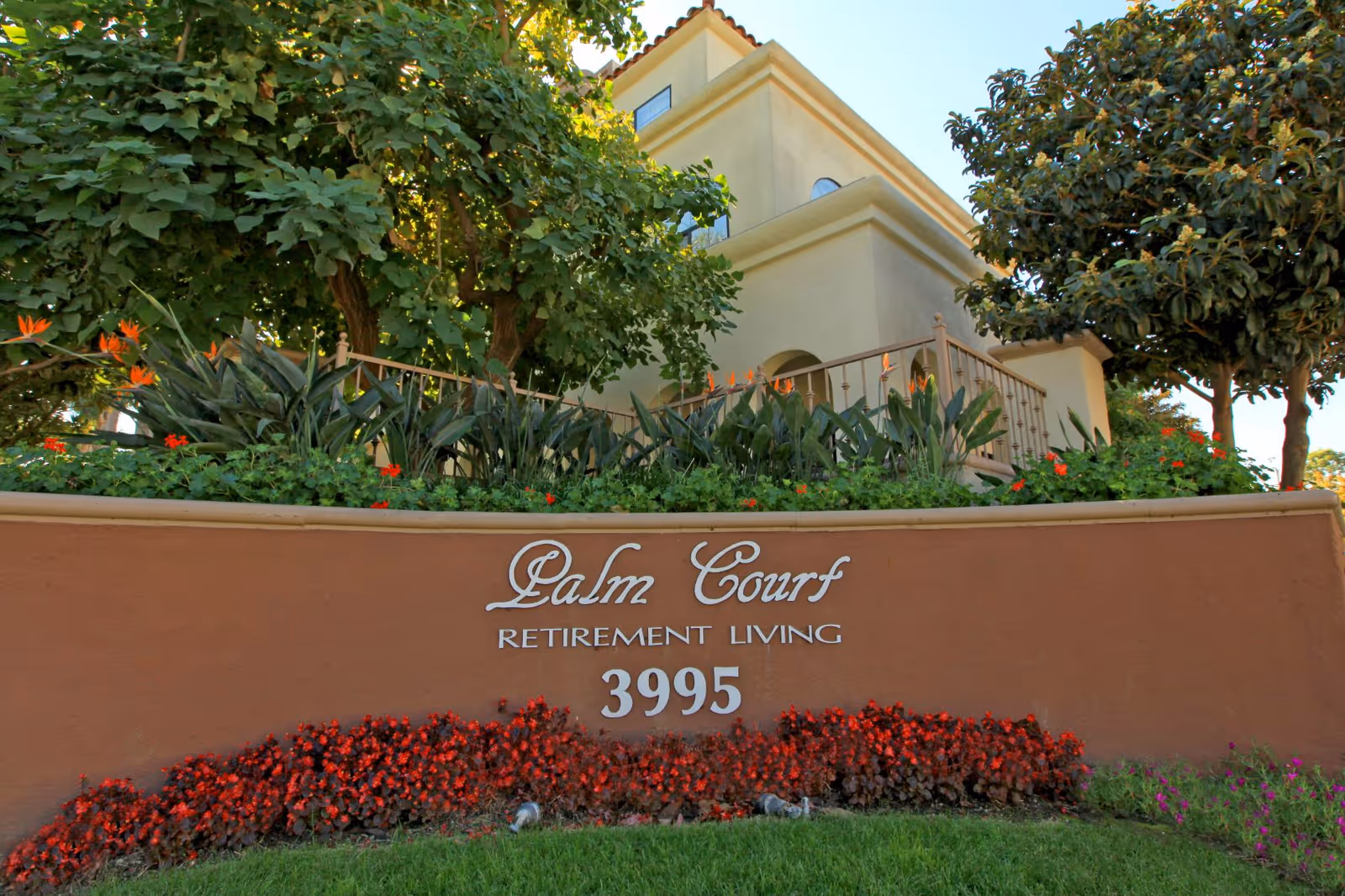 Entrance sign reading "Palm Court Retirement Living 3995" with landscaping and part of the building behind it.