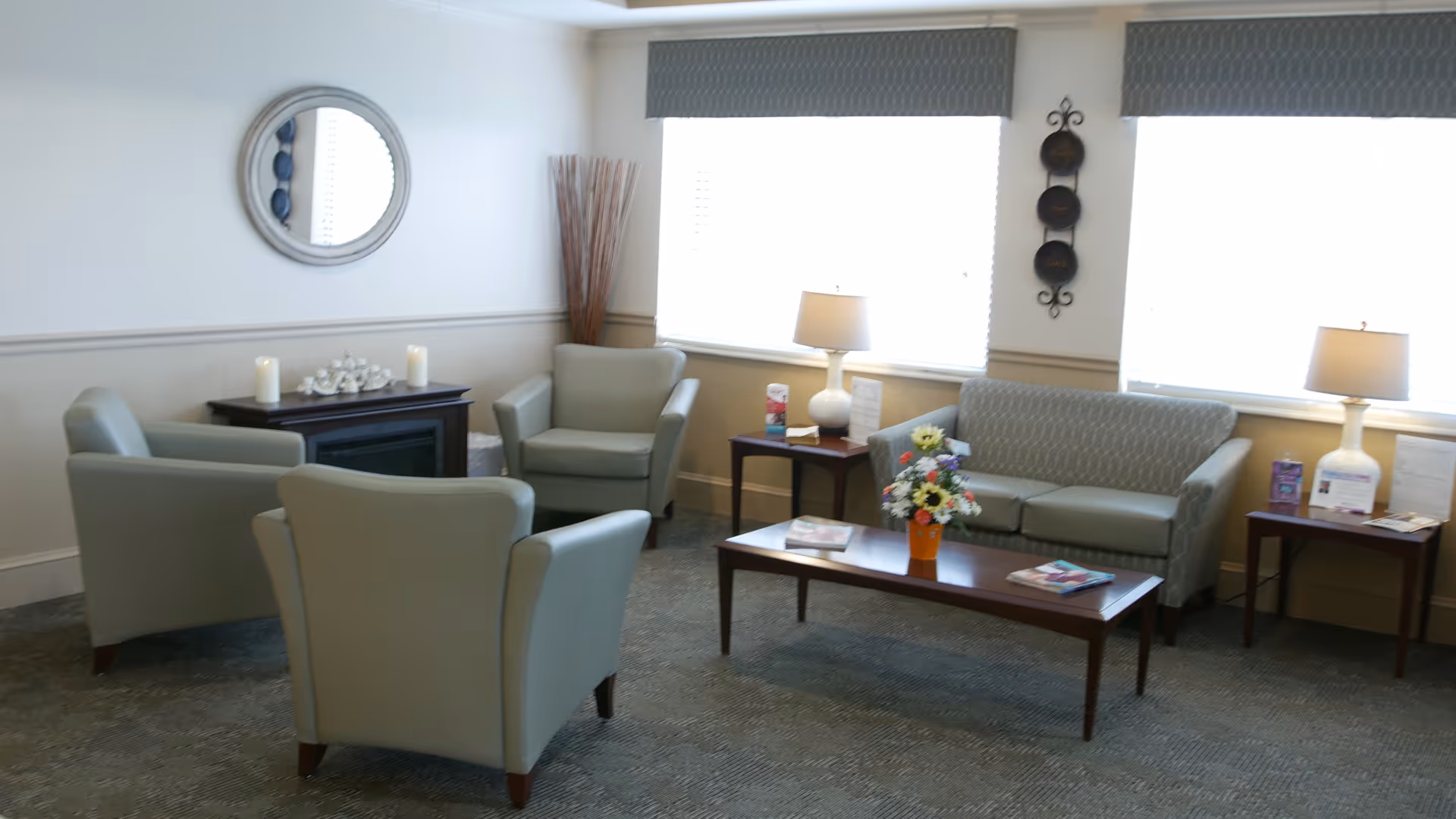 A cozy living room area in a senior living facility with light gray armchairs and a matching sofa arranged around a wooden coffee table with a flower vase and magazines. There are two side tables with lamps, a decorative mirror on the wall, and a small fireplace with candles and decor on top. Large windows with blinds allow natural light to fill the room.