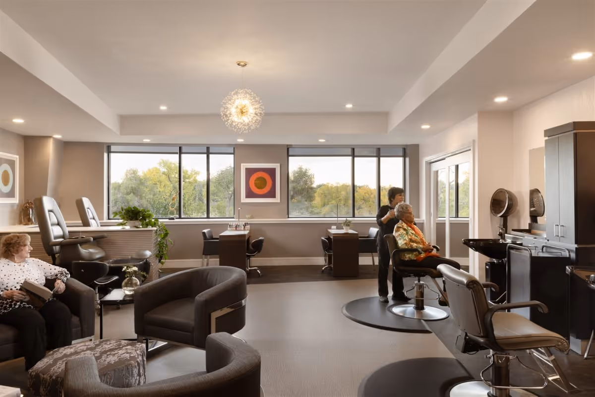 A bright and spacious salon area in a senior living facility with large windows showing greenery outside. The room features salon chairs and hair washing stations on the right, a woman getting her hair done by a stylist, and another elderly woman sitting on a couch reading a book. The decor includes modern furniture, plants, and framed artwork on the walls.