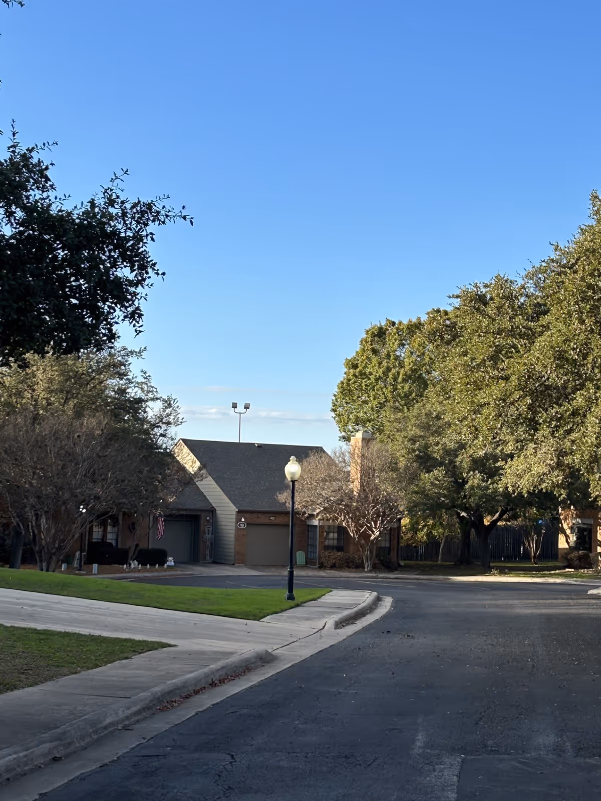 A quiet residential street within a senior living facility showing single-story brick homes with garages, surrounded by trees and green lawns under a clear blue sky.