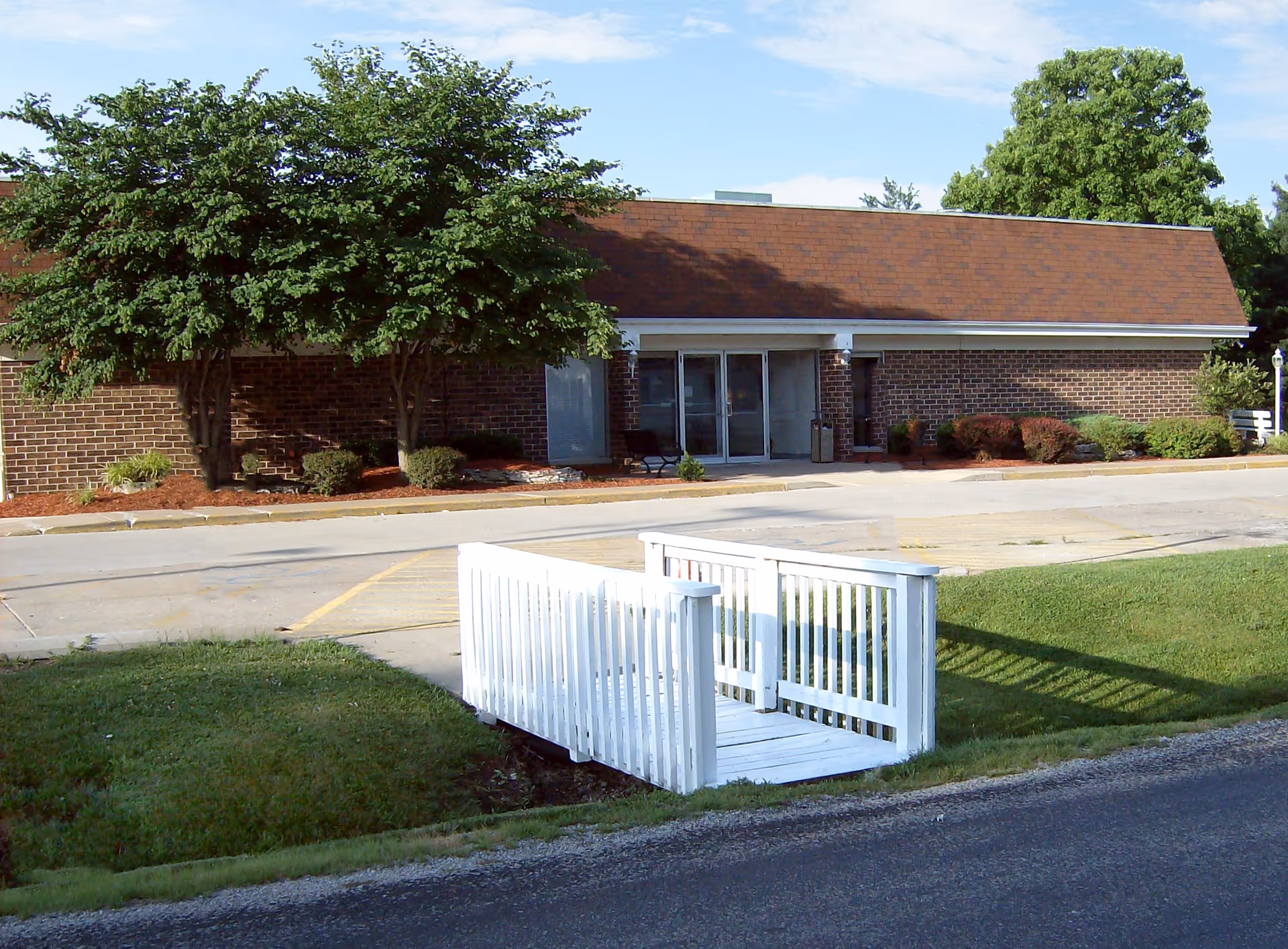 Exterior view of a single-story brick building with a brown shingled roof, glass double doors entrance, surrounded by landscaped bushes and trees. In the foreground, there is a small white wooden bridge over a grassy ditch leading to a paved parking area.