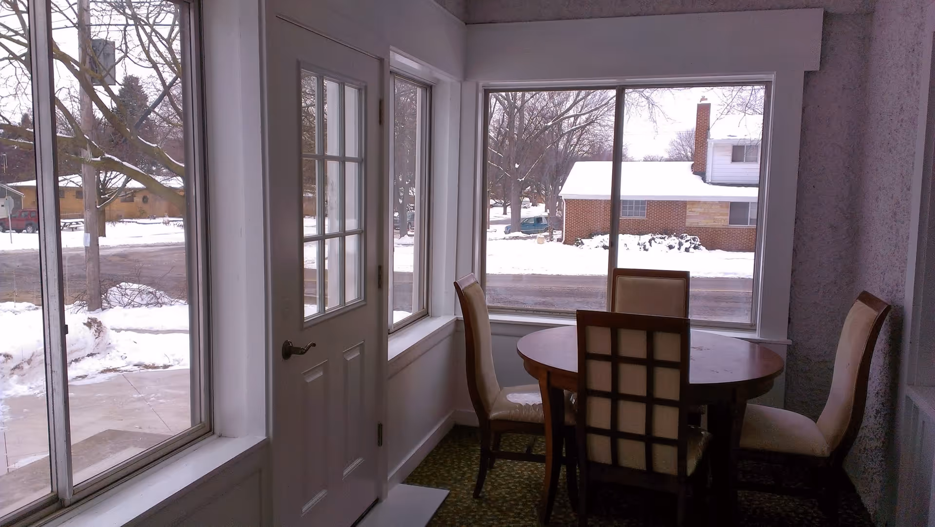 A small dining area with a round wooden table and four cushioned chairs near large windows showing a snowy outdoor scene with houses and trees.