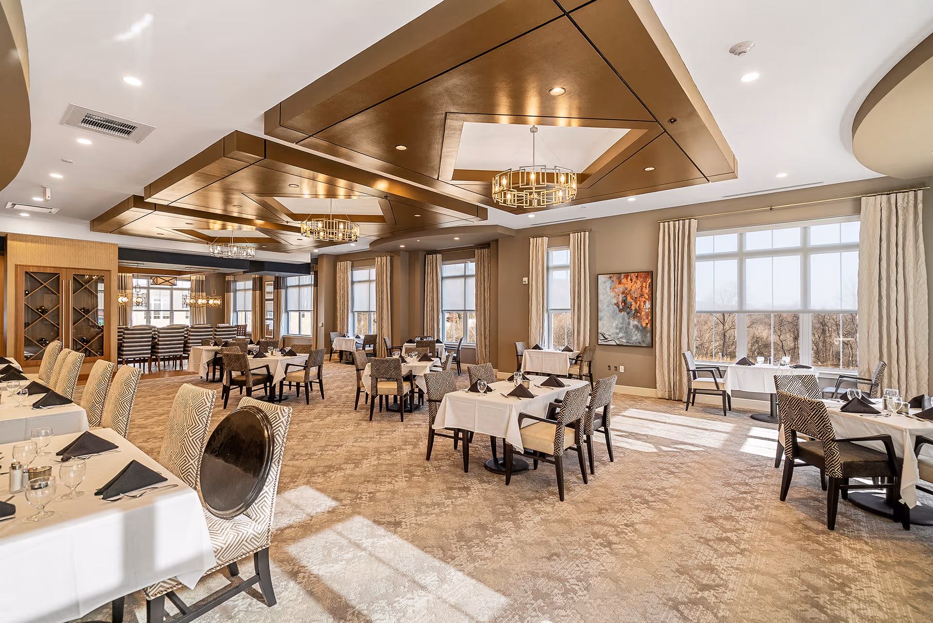 A spacious, well-lit dining room with multiple tables covered in white tablecloths, each set with glassware and black napkins. The room features large windows with beige curtains allowing natural light to fill the space, patterned carpet flooring, and decorative ceiling panels with modern chandeliers. Chairs have a mix of patterned and solid upholstery.