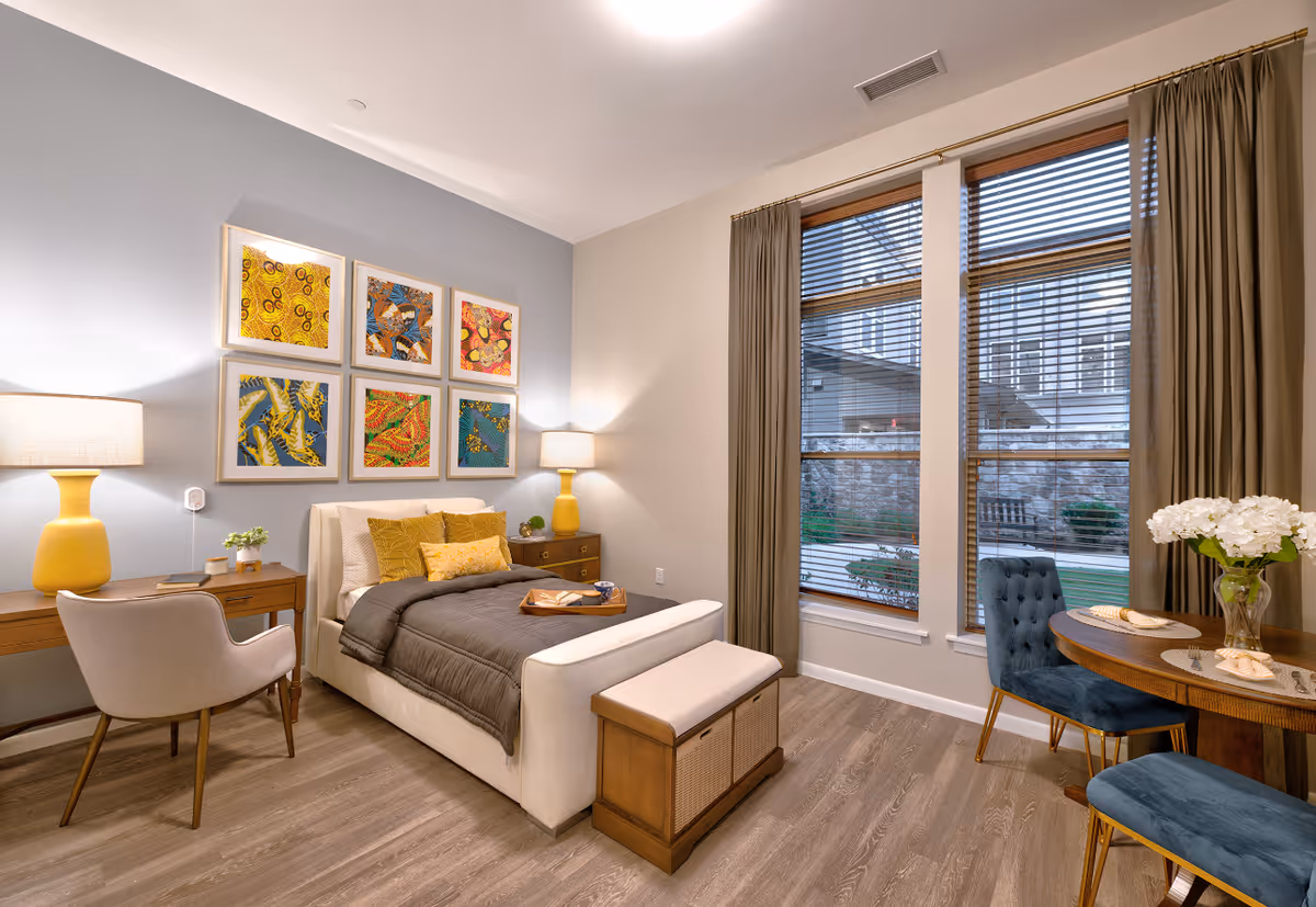 A cozy bedroom with a single bed featuring gray bedding and yellow pillows. The room has wooden flooring, a wooden desk with a beige chair and a yellow lamp on the left side, and a wooden nightstand with a matching yellow lamp on the right. Above the bed, there are six colorful framed artworks arranged in two rows. On the right side of the room, there are large windows with wooden blinds and brown curtains, next to a small round wooden table with two blue upholstered chairs and a vase of white flowers.