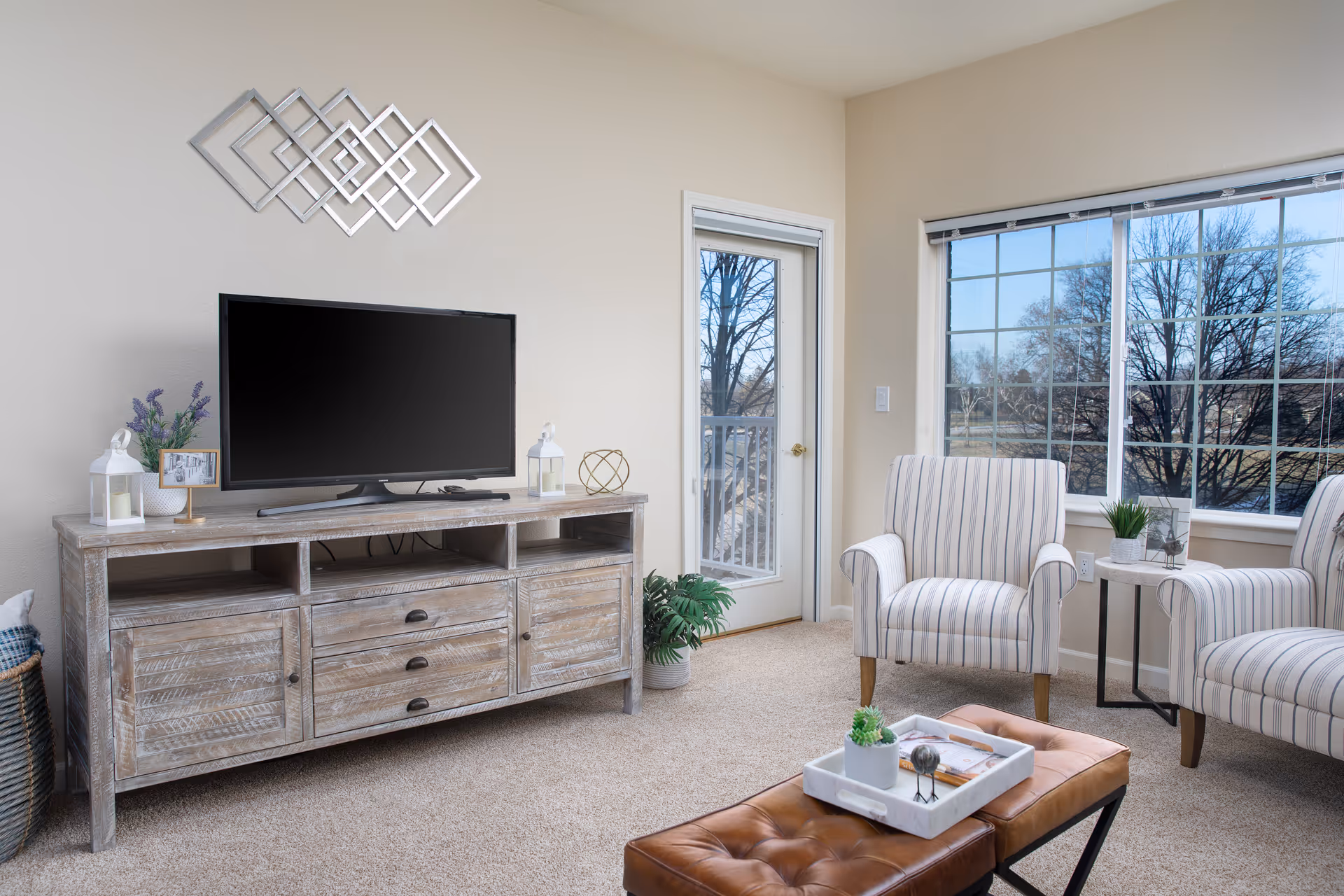 A bright living room with beige walls and carpet, featuring a wooden TV stand with a flat-screen TV, decorative items, and a basket on the side. Two striped armchairs are positioned near a large window and a glass door leading outside. A brown tufted bench with a white tray holding small plants and decor is in the foreground.