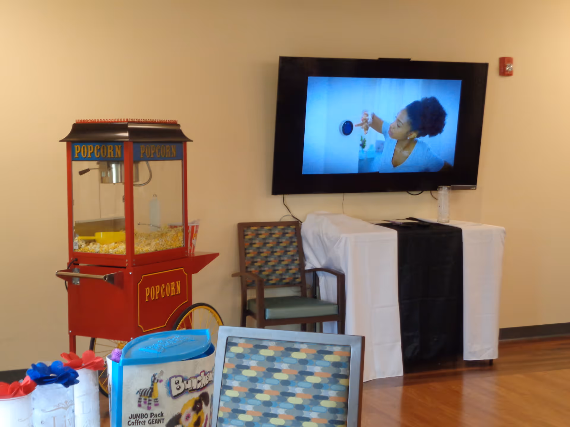 An indoor area with a red popcorn machine filled with popcorn on the left, a patterned chair, and a table covered with a white cloth and a black runner. A flat-screen TV mounted on the wall displays a woman interacting with a smart home device.