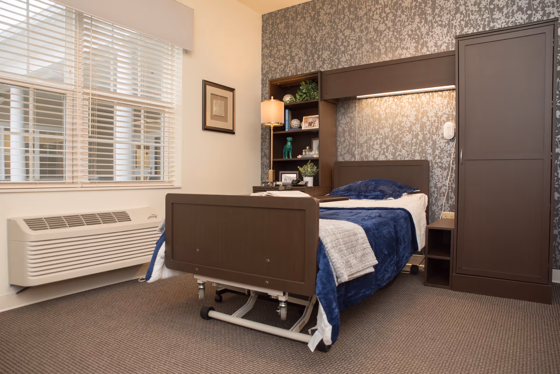 A furnished senior bedroom with a single adjustable bed, dark wood headboard and wardrobe, patterned wallpaper, and a window with blinds.