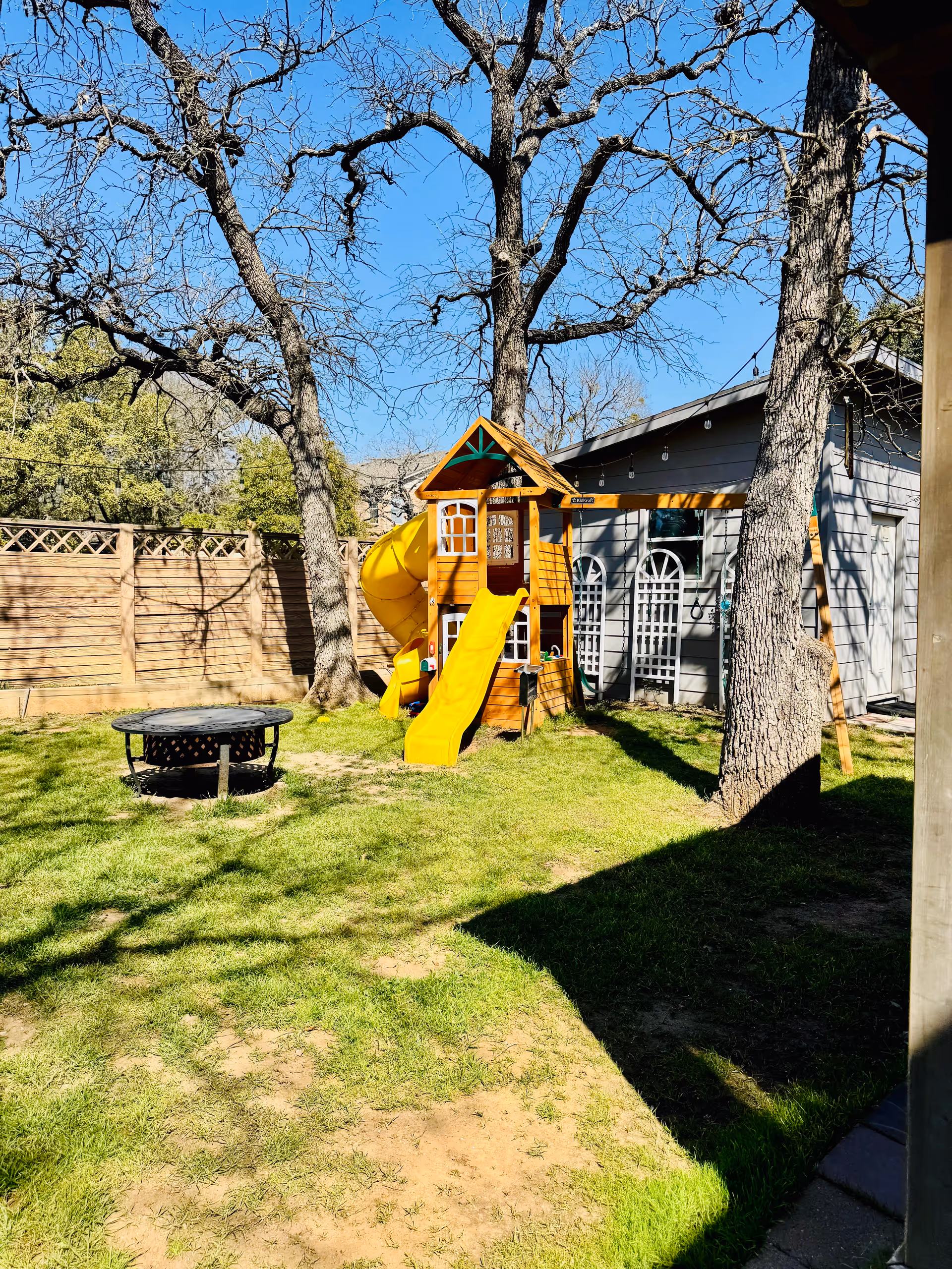 A sunny outdoor yard area with green grass, a wooden play structure featuring a yellow slide and a spiral tube slide, surrounded by leafless trees and a wooden fence. There is a small fire pit on the grass and a gray building with arched windows in the background.