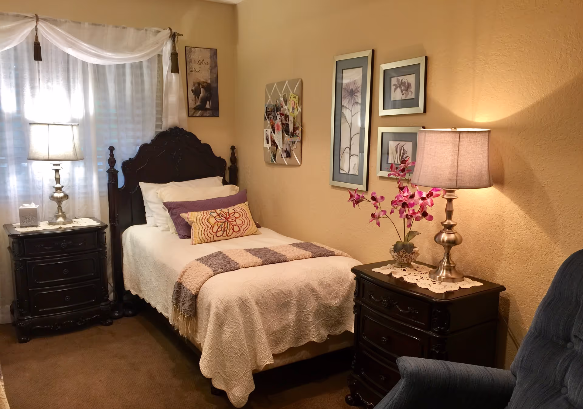 A cozy bedroom with a single dark wooden bed featuring white and purple bedding. There are two dark wooden nightstands on either side of the bed, each with a lamp. One nightstand has a tissue box, and the other has a vase with purple flowers. The walls are beige with framed floral artwork and a bulletin board with photos. A blue upholstered chair is partially visible in the foreground.