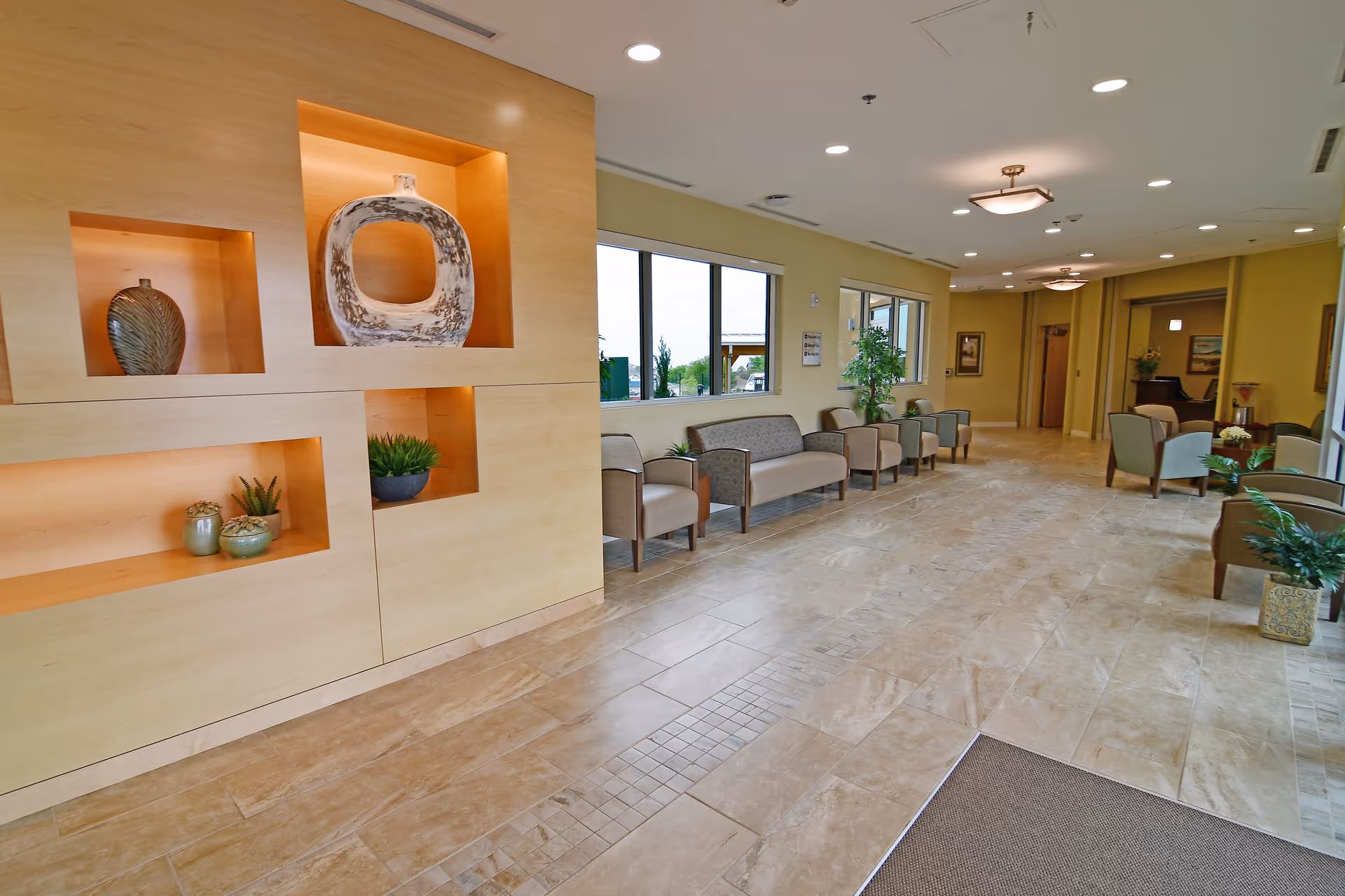 A spacious and well-lit interior common area with beige tiled floors and light yellow walls. The room features a built-in wooden display with decorative vases and plants, several chairs and sofas arranged along the walls, large windows letting in natural light, and ceiling lights providing additional illumination.