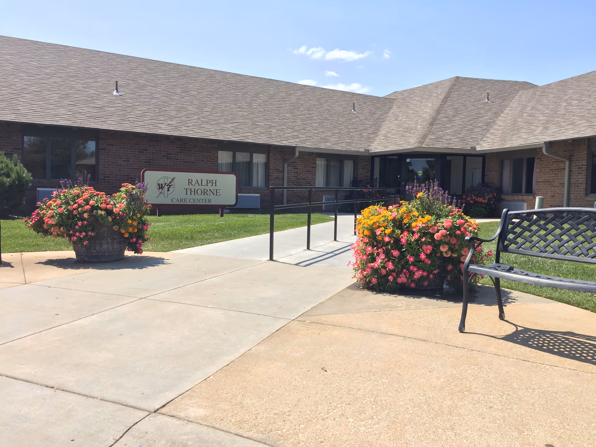 Exterior view of the Ralph Thorne Care Center building with a paved walkway leading to the entrance. There are large flower pots with colorful flowers on either side of the walkway and a black metal bench on the right side. The building has a brick facade and a sloped roof under a clear blue sky.