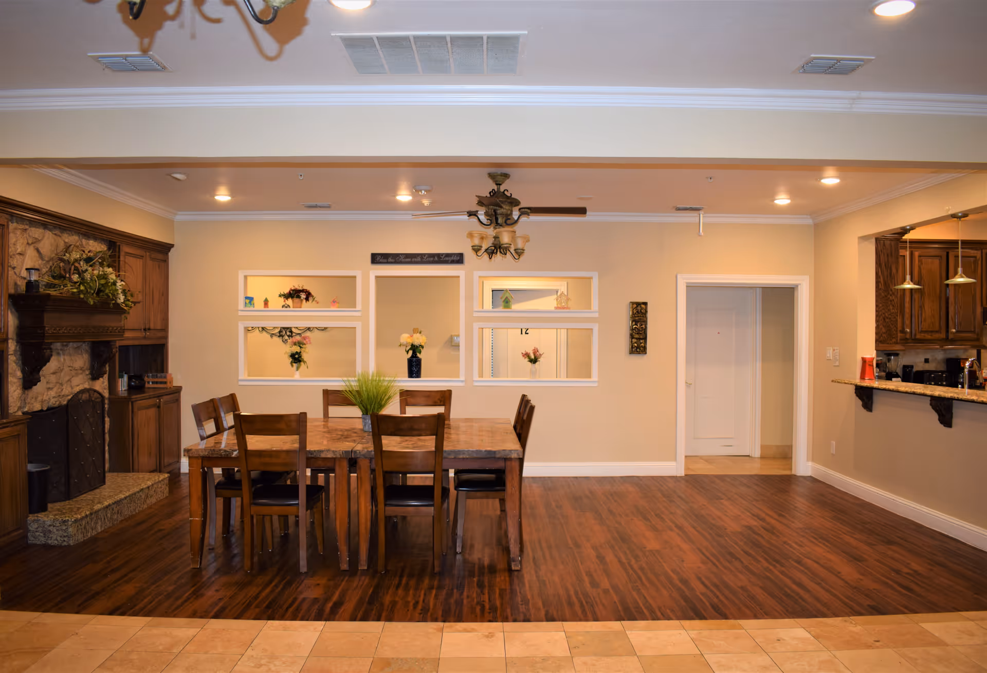 Interior view of a dining area in Avalon Memory Care facility featuring a wooden dining table with six chairs on a dark wood floor. To the left is a stone fireplace with a wooden mantel and built-in cabinets. The back wall has decorative windows with flower arrangements and a sign above them. To the right is a kitchen counter with hanging pendant lights and wooden cabinets.