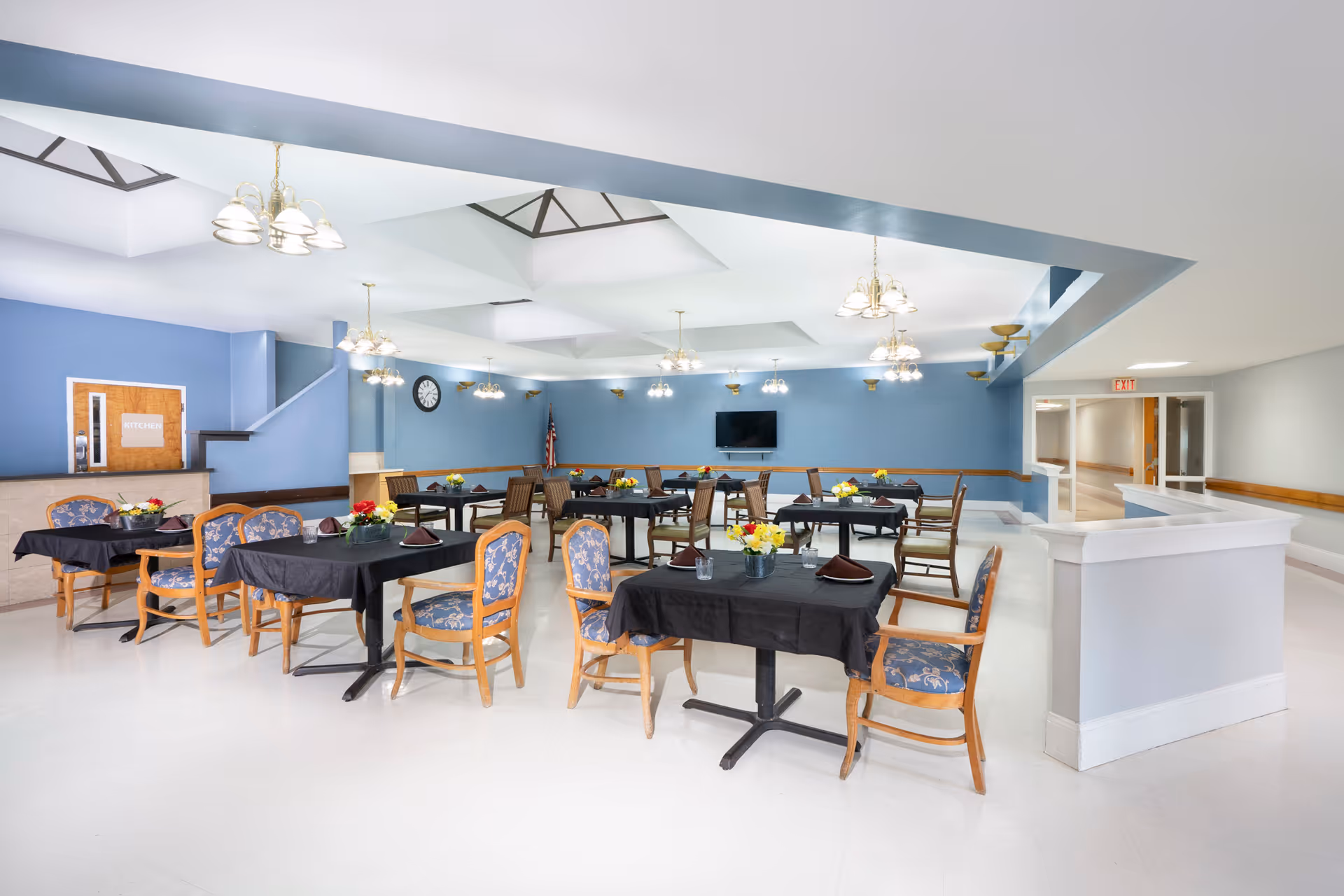 A spacious dining room with multiple tables covered in black tablecloths, each set with chairs featuring blue floral upholstery. The room has light blue walls, several ceiling light fixtures, a wall clock, a mounted television, and an American flag in the corner. A door labeled 'KITCHEN' is visible on the left side of the room.