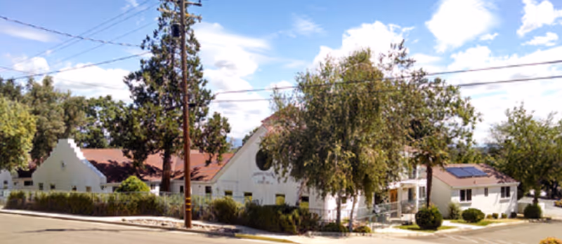 White single-story assisted living building with red roofs, trees, and power lines viewed from the street under a blue sky.