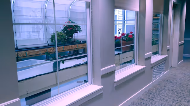 Interior hallway with large windows looking out onto an enclosed garden area with planter boxes containing red flowers. The hallway has light-colored walls and carpeted flooring.