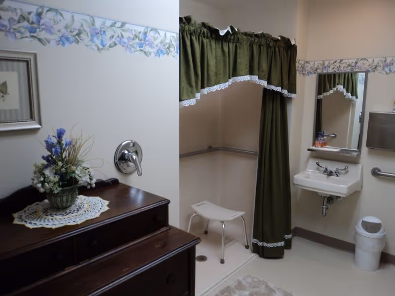 A bathroom with a walk-in shower area that has a green curtain with white lace trim and a white shower stool. There is a small white sink with a mirror above it, a soap dispenser, a trash can, and a wooden dresser with a floral arrangement on top. The walls have a floral wallpaper border near the ceiling.