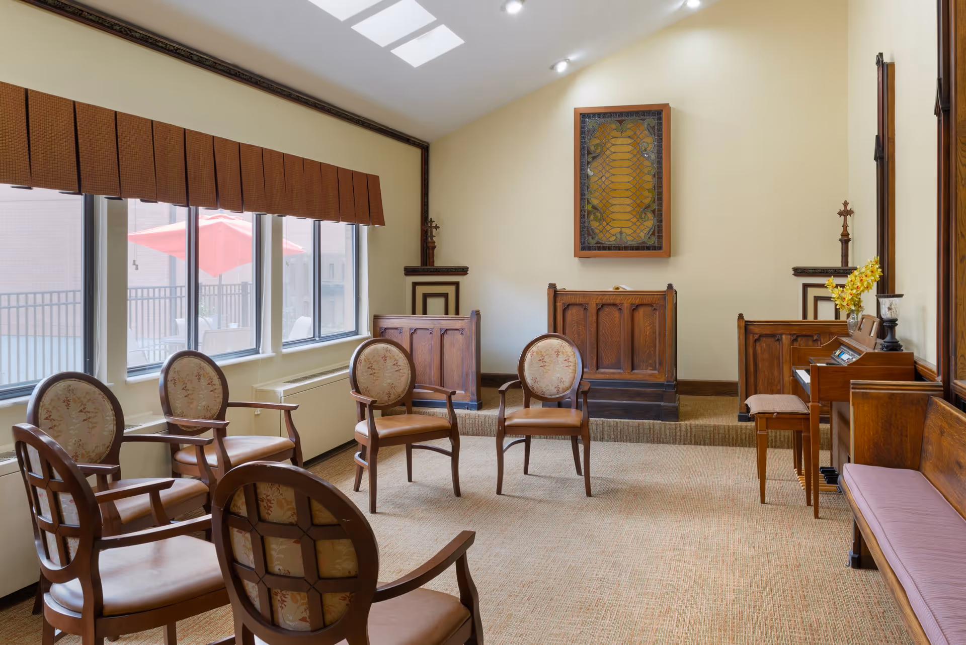 Sunlit interior meeting/chapel room with arranged wooden chairs, a wooden podium and decorative wall art beside large windows.